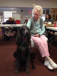 Pictured is an older woman with a black poodle as her service dog companion.